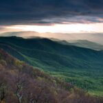 Forest covered mountains with clouds overhead and sun just peeking out in between.