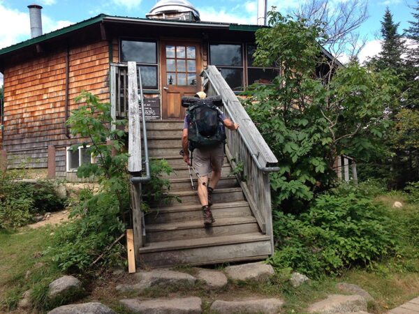 Hiker wearing pack climbs short staircase into a small wooden building