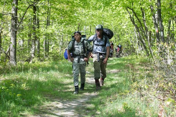 Two teenage hikers walk along a trail, with one gently resting a hand on the other's back in support.