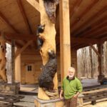 Todd Gladfelther wearing a green sweatshirt and brown pants, standing next to his carving of bears climbing up a post.