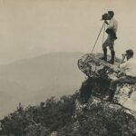 Seipa photo of George Masa standing on a rocky outcropping surrounded by vegetation taking a photo from Graybeard mountain, accompanied by Horace Kephart.