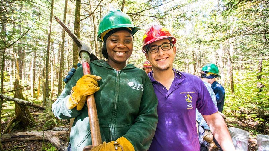 Two Trail Crew members smile side by side wearing helmets and holding tools.