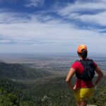 Trail runner wearing an orange hat, red shirt, yellow shorts, and lightweight running vest looking out at the scenic landscape.