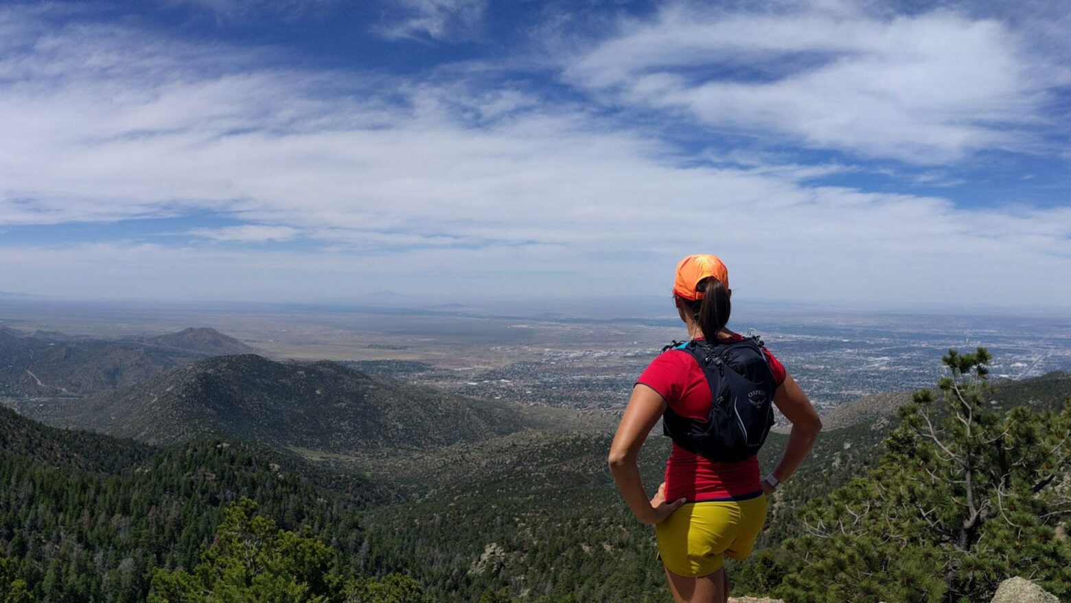 Trail runner wearing an orange hat, athletic red shirt and yellow shorts, and lightweight running vest looking out at the scenic landscape.