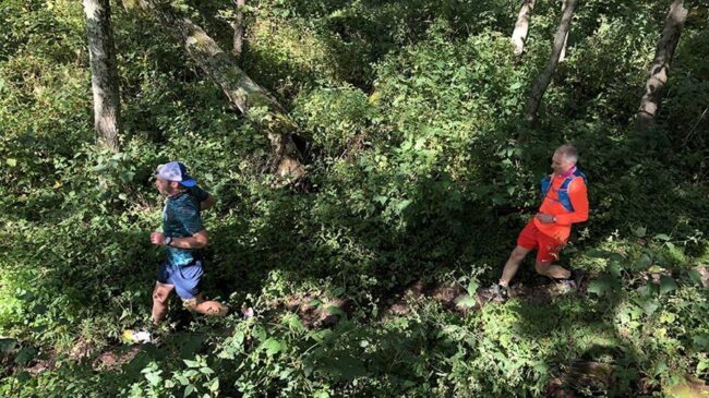 Two runners jog along a green, single-track