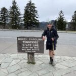 Hiker holding hiking poles and posing next to wooden sign reading "North Carolina Tennessee State Line"