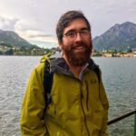 Portrait of an adult man with dark hair, a beard, and glasses. He wears a chartreuse jacket and stands in front of a lake with mountains in the background.