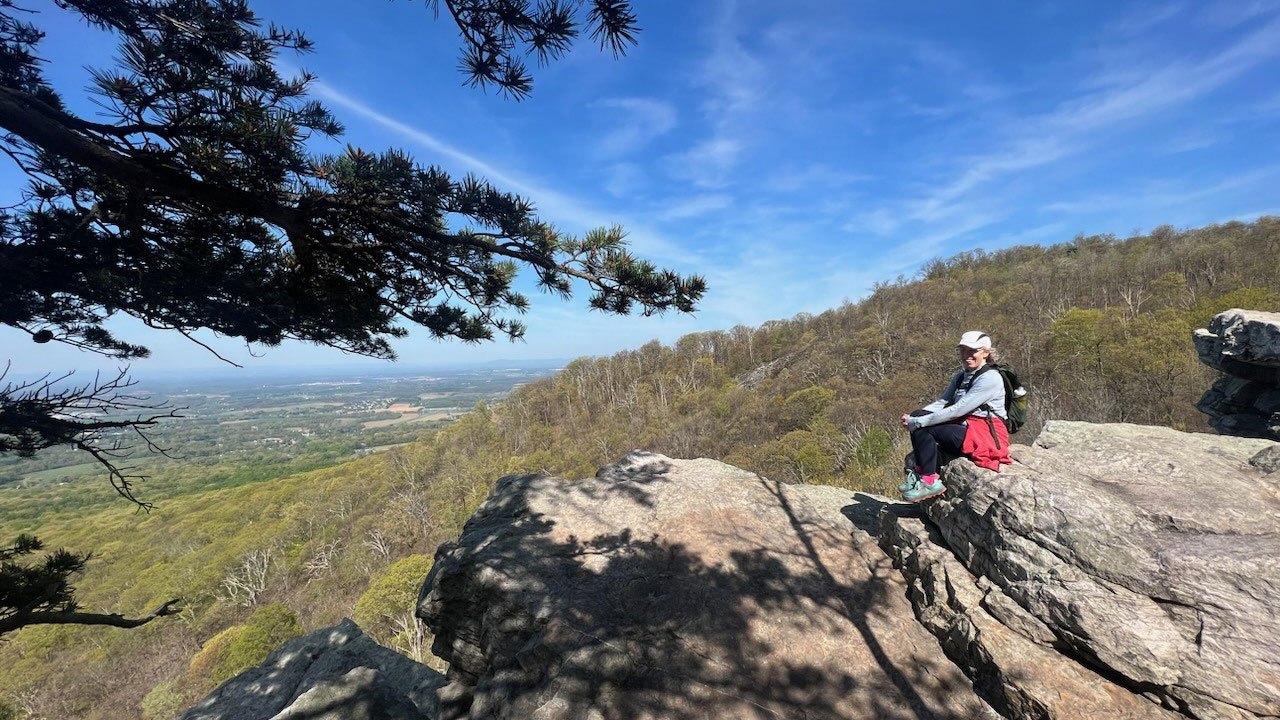 A female hiker sits in the sun at Annapolis Rock and smiles toward the camera