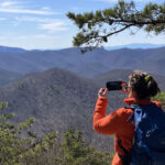 Woman wearing an orange jacket and blue pack, holding up her smartphone to capture a photo of the mountains in the distance.
