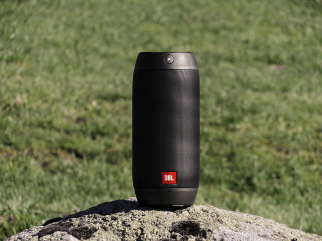 A small, black portable speaker sits on a rock with green grass in the background