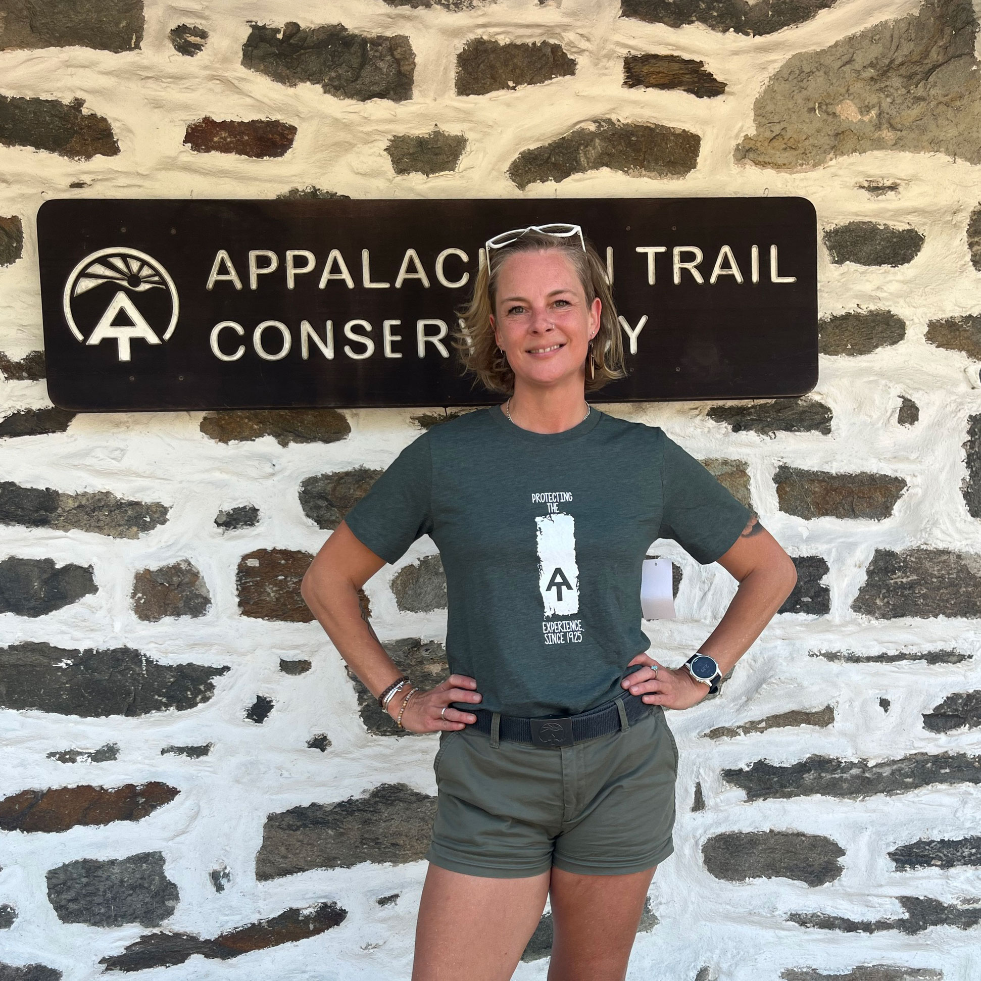 Woman wears an olive green ATC t-shirt with a white blaze and A.T. symbol on the front