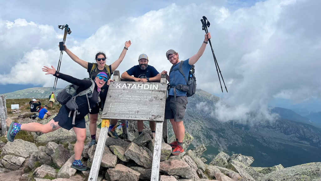 A group of four hikers pose with the Katahdin summit sign, celebrating their completed thru-hike.