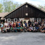 Photo of 33 people wearing outdoors clothing, smiling and posed outside the front of a building with a nameplate reading "Big Timbers"