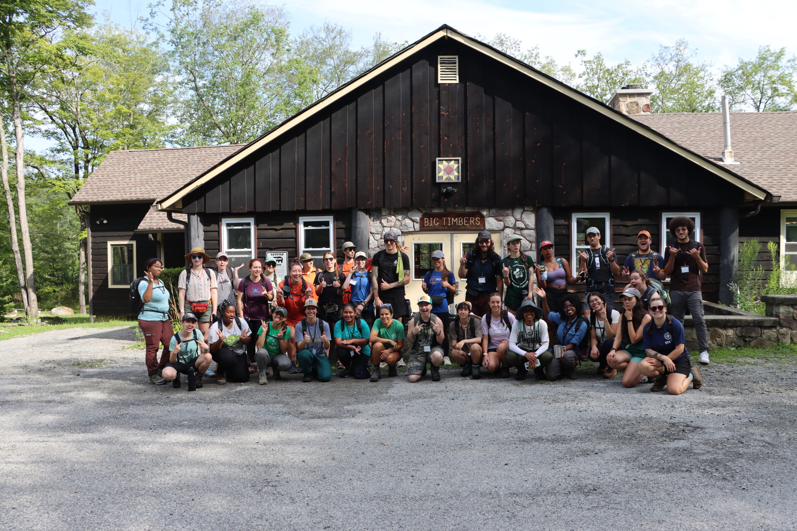 Photo of 33 people wearing outdoors clothing, smiling and posed outside the front of a building with a nameplate reading "Big Timbers"