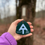 2025 hiker hangtag held in front of a tree marked with a white blaze.