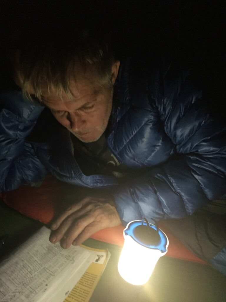 Bob "Blue" Parker wearing a blue puffy jacket and lying on a red sleeping pad while reading a book in the dark by the light of a lantern.