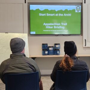 Two hikers watch a presentation with the slide title "Start Smart at the Arch!"