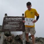 A photo of A.T. volunteer Ron Hudnell holding a sign the says "RLTW on the A.T." next to a sign on Mount Katahdin.