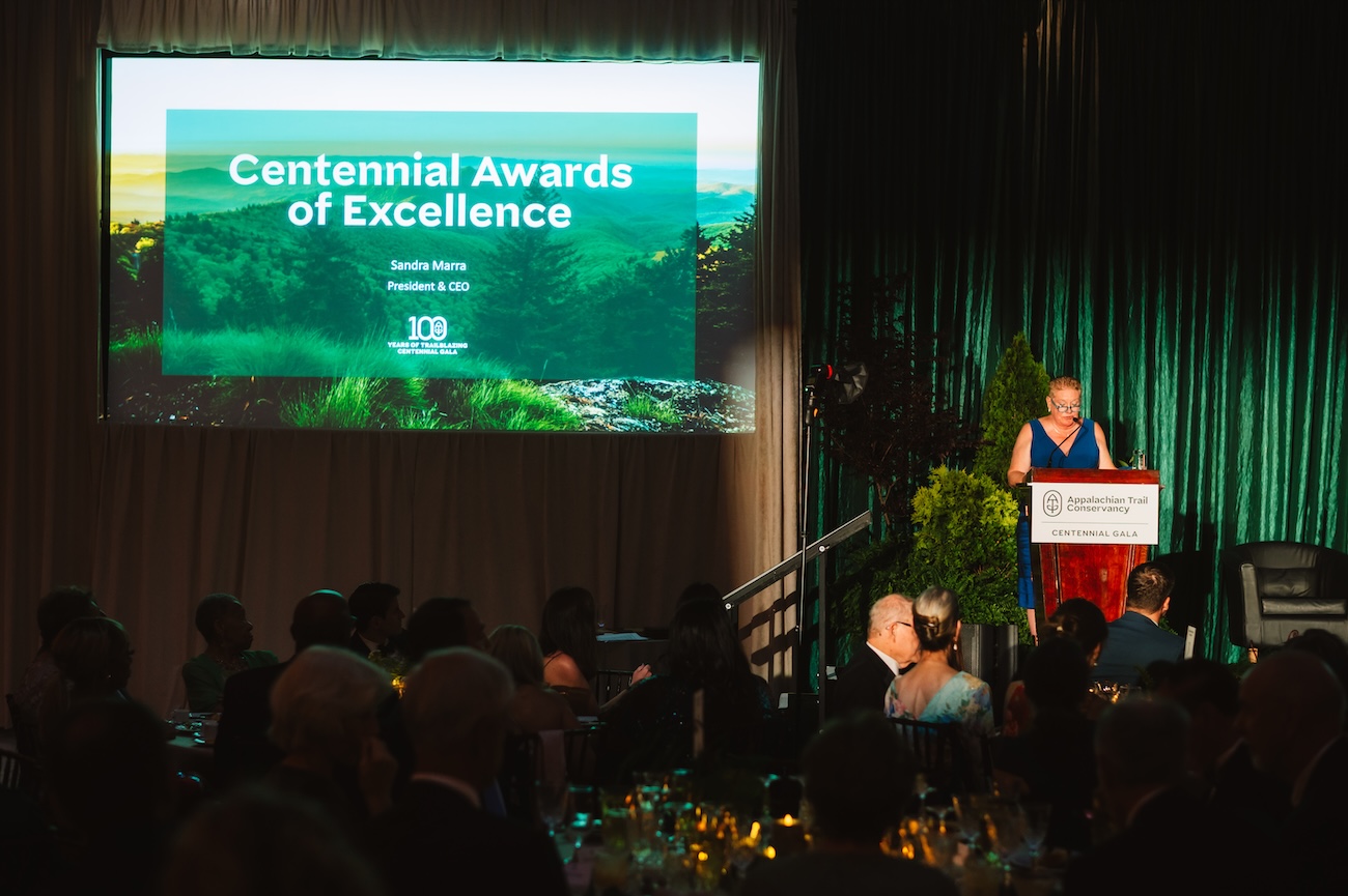 ATC President and CEO Sandi Marra speaks at a lit podium, addressing the audience. Behind her, a large screen displays the words "Centennial Awards of Excellence."