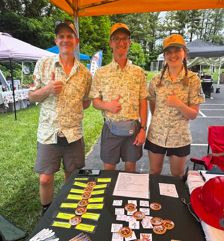 Three PATC Ridgerunners wearing matching shirts smile and give a thumbs-up to the camera while standing behind a table covered with PATC buttons and informational materials.