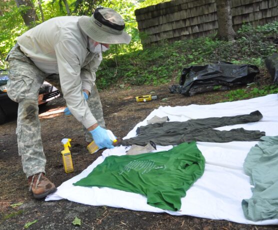 Person wearing a mask sprays permethrin onto clothing spread on the ground.
