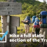 Five hikers hike past Grayson Highlands State Park sign on the Appalachian Trail. Text overlay "Hike a full state section of the Trail" next to a checkbox.