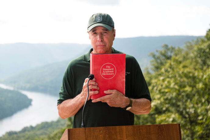 Kevin Nealon stands at a podium wearing an ATC polo shirt and hat, holding up a dictionary with a river and mountain landscape in the background.