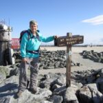 Hiker poses next to wooden sign reading "Mount Washington Summit, 6,288 feet / 1,917 meters"