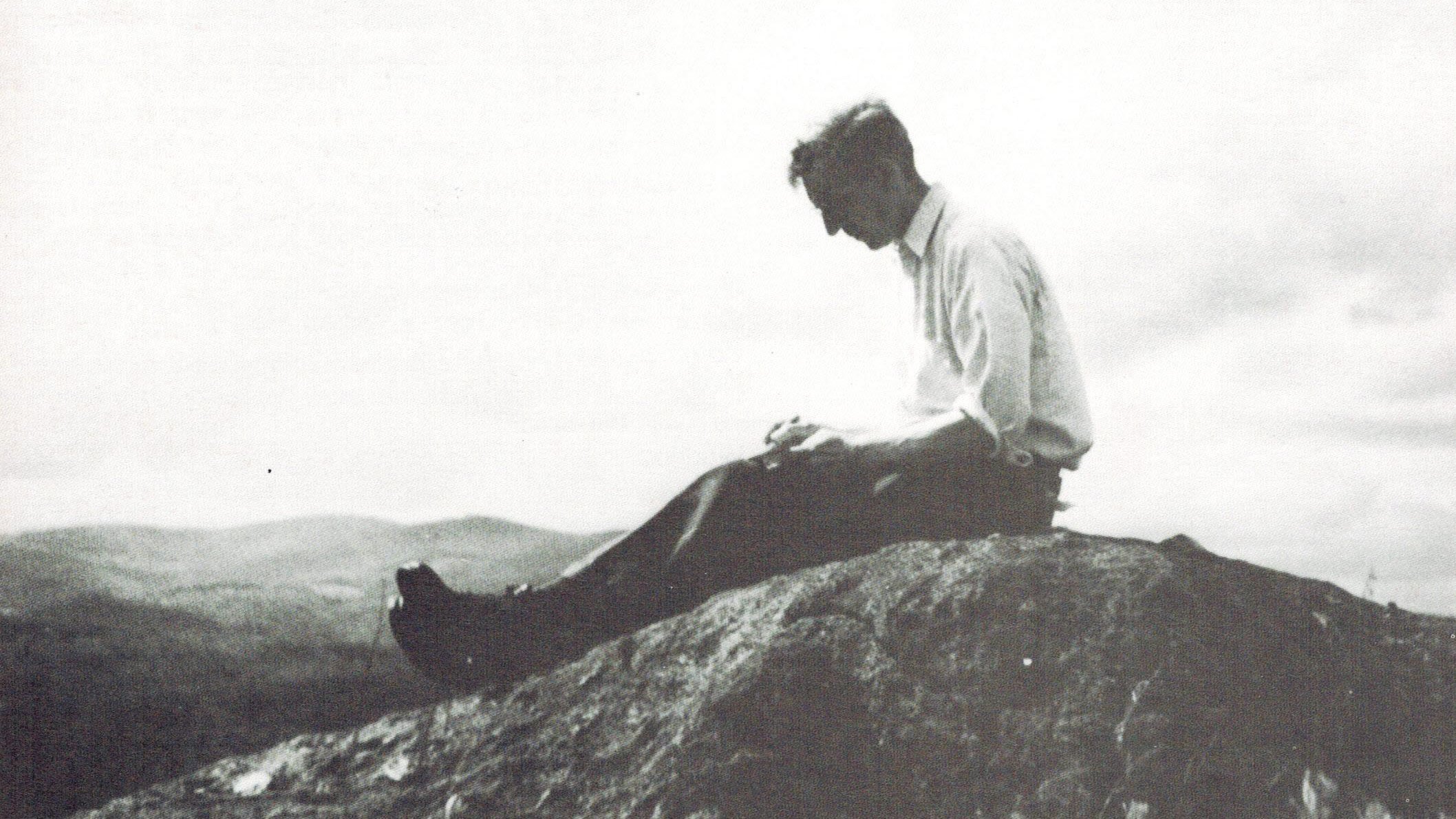 Benton MacKaye in a black-and-white photograph sitting atop a rocky overlook, looking down at a notebook.