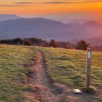 White blaze painted on a wooden trail marker at Max Patch, set against the grassy bald and a vivid orange sunset in the background.
