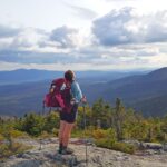 Female hiker with a backpack and trekking poles stands with arms outstretched, facing a mountain landscape.