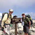 Two hikers smile and pose next to Mount Washington Summit sign