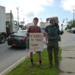 Two hikers stand by the roadside. One holds a sign that says “A.T. Hiker to Trail, Clean, Just Showered,” while the other jokes by smelling their armpit.