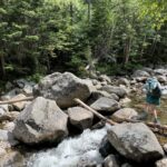 Hiker using trekking poles and a fallen log to cross a rocky stream.