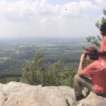 Two Soul Trak hikers enjoying a scenic farmland overlook, with one using binoculars to observe a bird.