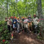 Volunteers in hard hats celebrating as they cut a ceremonial ribbon of pink flagging tape using loppers.