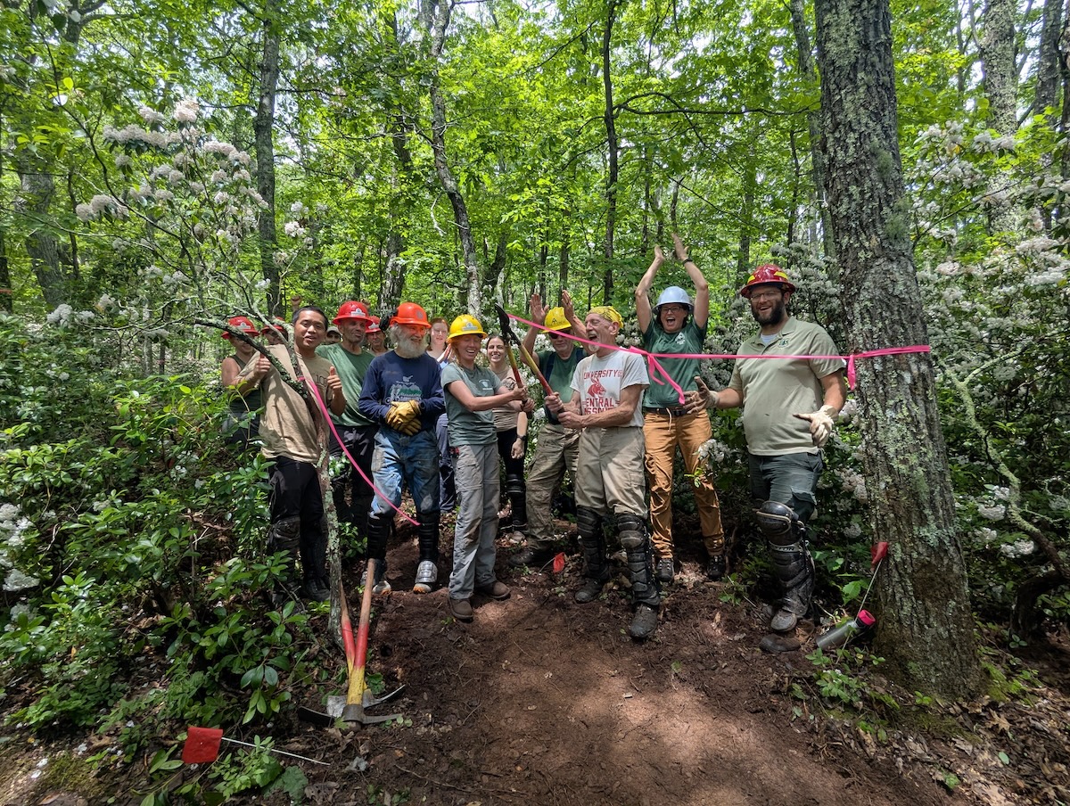 Volunteers in hard hats celebrating as they cut a ceremonial ribbon of pink flagging tape using loppers.