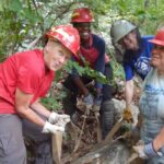 Four Appalachain Trail volunteers wearing hardhats and gloves use straps to lift a large rock.