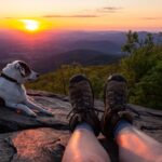View of hiker's boots and leashed dog with a scenic mountain vista and sunset in background.