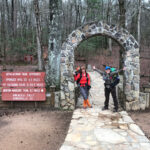 Two hikers pose under a stone archway marked "Appalachian Trail Approach"
