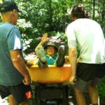 Inch Worm laughs and waves at the camera as two hikers carry her in a wheelbarrow along the Trail.
