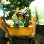 Inch Worm laughs and waves at the camera as two hikers carry her in a wheelbarrow along the Trail.