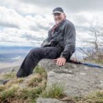 The Scotsman sits on a rocky overlook with hiking poles at his side.