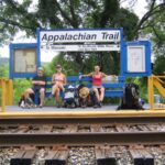 Three hikers rest on a blue bench at an Appalachian Trail trailhead sign by the tracks, with backpacks on the platform.