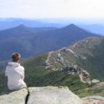 Person in a white jacket sits on a rocky ledge overlooking a vast, forested mountain range.