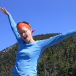 Young woman in a blue long-sleeve shirt and orange bandana with arms raised in a joyful pose against a clear blue sky and forested hills behind her, wearing glasses.