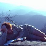 Man lying on a rocky mountaintop, propped on one arm and reading a book with a pencil in his mouth, wearing a light jacket. The background shows blue sky and distant mountains.