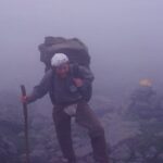Hiker with large backpack and trekking pole navigating a foggy rocky mountain trail.