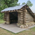 A three-sided shelter made of wood, stone, and a tin roof positioned on a raised concrete slab for display.
