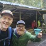 Two hikers take a selfie in a forest, with a wooden shelter behind them and a teal cup in the foreground.